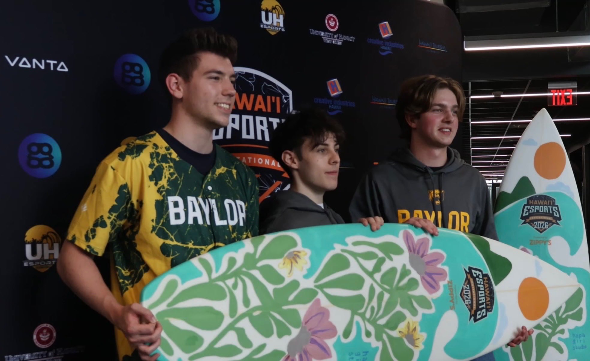 Three Baylor Esports team members stand together in front of a Hawai‘i Esports Invitational backdrop, holding surfboard trophy.