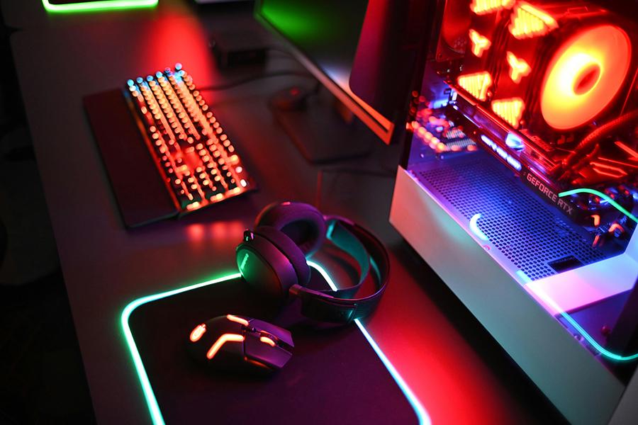 RGB-lit gaming keyboard, mouse, and headphones on a glowing mouse pad, with a desktop PC showing red and blue LED lights.